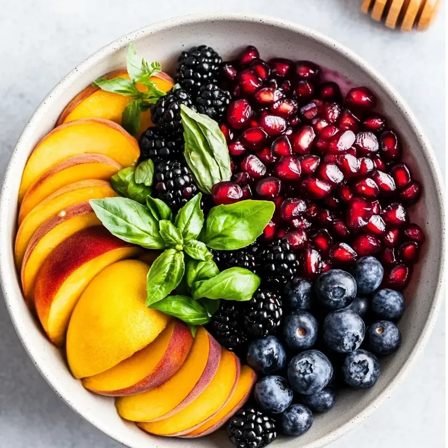 A bowl of fruit with blueberries, blackberries, peaches, and honey.